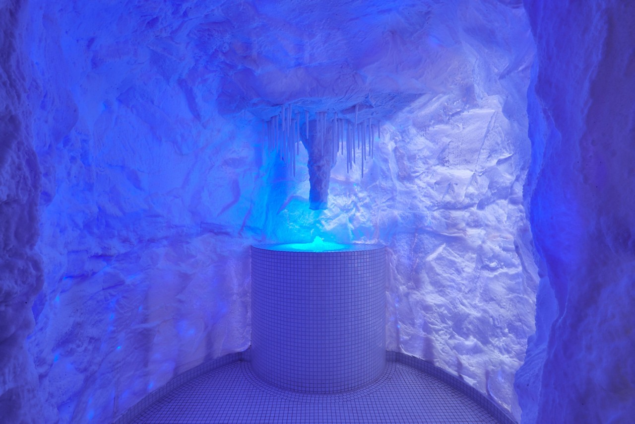 Cylindrical tiled basin glows blue while water or light pours from hanging icicles above, set within a cave-like, ice-themed room with textured white walls and curved tiled floor.